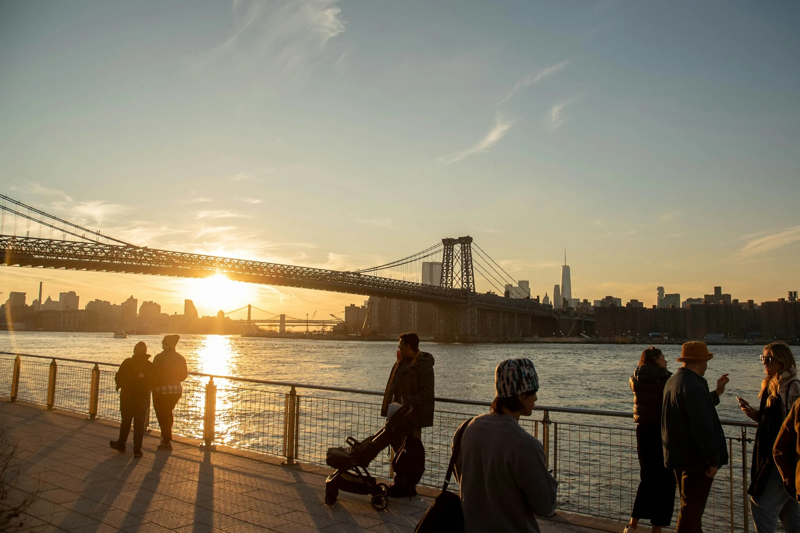 Golden evening light across the East River, with a steel bridge, the Manhattan skyline and people along the Brooklyn waterfront.